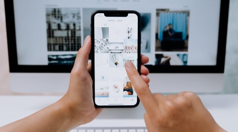 Hands holding a smartphone while browsing a social media app in front of a desktop computer.