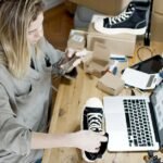 Woman photographing shoes for online sale in home workspace with laptop and packages.